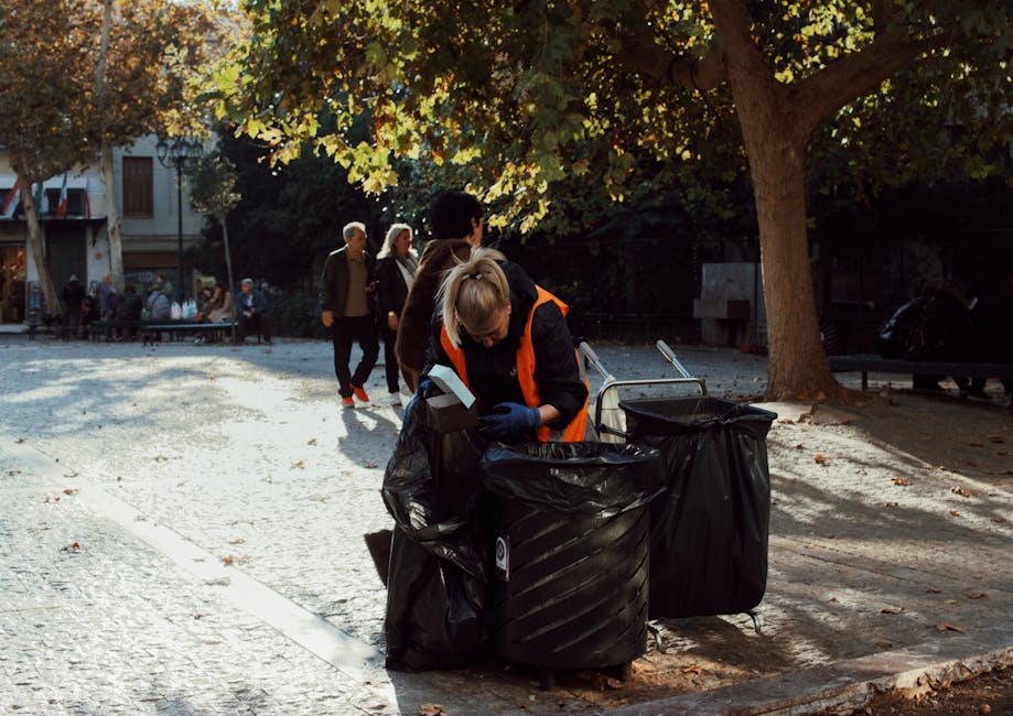 A woman wearing a high-visibility orange and black jacket is bending over to collect waste or recyclables from a black plastic rubbish bag placed inside a large outdoor bin situated on a paved surface. The scene is set in a park or urban public space shaded by a large tree with green and yellow leaves, indicating autumn. In the background, several other individuals, some walking and others standing, are visible, along with a building and additional trees. The sunlight filters through the foliage, creating shadows on the ground. The overall environment appears calm and organized, with the activity of waste collection or disposal which aligns with services offered by Waste Clearance Tufnell Park for rubbish removal and private waste handling.
