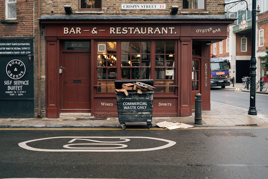 In front of a red-brick building with large windows, a black commercial waste bin labeled 'Commercial Waste Only' is positioned on the sidewalk, filled with flattened cardboard boxes and paper debris. The bin is situated near a small grey bollard and a metal post, with some paper and cardboard lying on the pavement nearby. The storefront appears to be a bar and restaurant, with signage indicating wine, spirits, and oyster bar offerings, and a smaller blackboard sign advertising a self-service buffet. The street features a marked parking area with a white outlined parking symbol painted on the tarmac. In the background, a silver truck with a blue cab is driving past, and a bicycle is parked further down the sidewalk. The scene suggests an urban environment where external waste collection services, such as those offered by Waste Clearance Tufnell Park, are used for managing waste generated by local businesses, supporting alternative waste handling solutions outside of municipal collection.
