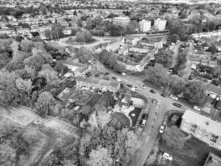 An aerial black-and-white photograph of a residential area showing a mix of houses, apartment buildings, and surrounding trees. In the foreground, there is a collection of small, single-story houses with sloped roofs and neatly maintained gardens, some with fencing and patio areas. The street runs centrally through the scene, with multiple parked cars on both sides, and a few vehicles in motion. To the left, a large area of open space or parking lot with marked parking bays is partially visible, bordered by trees. In the background, there are several multi-story buildings, likely apartments or offices, along with tree-lined streets and patches of greenery, giving a sense of a well-developed local neighbourhood. The scene highlights a typical urban setting where private property and public roads coexist, illustrating a context suitable for services involving rubbish clearance or alternative waste handling managed by companies such as Waste Clearance Tufnell Park.