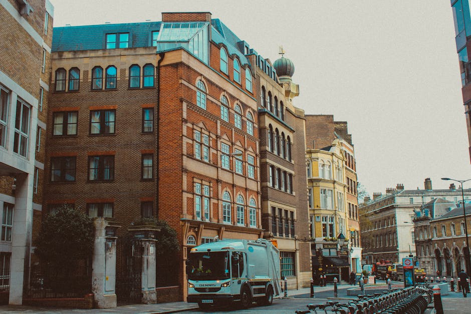 A city street scene featuring a collection of historic and modern buildings with various architectural styles, including red brick facades, arched windows, and decorative cornices, situated along the pavement. In the foreground, a waste collection vehicle with a light blue body and black wheels is parked close to the curb, next to a low stone fence and some greenery. The street is lined with metal bollards and bike racks, with additional vehicles and pedestrians visible in the background. The environment appears to be in an urban area with a mix of commercial and residential structures, illuminated by natural daylight with overcast skies. This scene exemplifies an area where private waste removal services like Waste Clearance Tufnell Park may operate, demonstrating typical street-level activity and infrastructure related to rubbish management in a busy city setting.