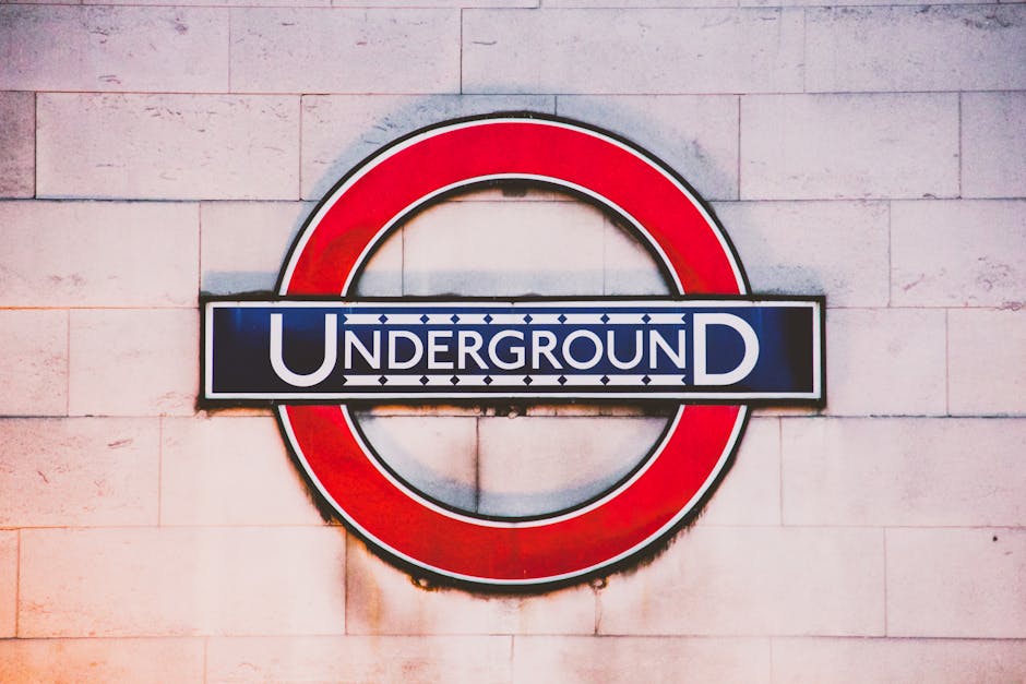 A close-up image of a London Underground station sign mounted on a light-colored brick wall. The sign features a horizontal black rectangular background with the word 'UNDERGROUND' in white capital letters, and a prominent red circular border surrounding the sign. The letters 'U' and 'D' are larger, with 'UNDERGROUND' written across the center, creating a clear focal point. The wall has visible texture with slight discoloration and subtle marks, indicating an exterior or well-worn interior setting. The positioning of the sign suggests it is displayed at an accessible height for travelers, contributing to the station’s functional appearance. While the sign itself is not directly related to rubbish removal, the clean, clear presentation of the signage and wall surface reflects typical urban environments that Waste Clearance Tufnell Park might service for maintenance or cleaning tasks in areas requiring regular rubbish clearance, especially around public transportation hubs where waste accumulates from daily foot traffic.