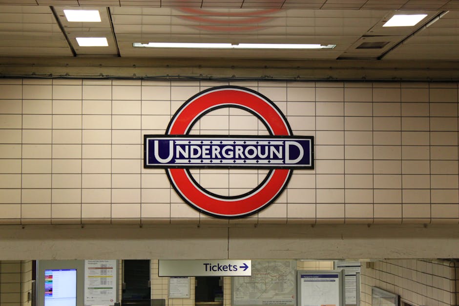 A large circular London Underground sign is mounted on a beige tiled wall in an underground station. The sign features a red outer circle with a black border and a blue rectangular bar across the middle displaying the word 'UNDERGROUND' in white capital letters, with a black and white dotted pattern behind the text. Below the sign, there is a white directional sign with the word 'Tickets' and an arrow pointing to the right, indicating the location of ticketing facilities. The ceiling above consists of white panels with embedded fluorescent lighting, creating a well-lit environment. The overall setting is clean and organized, typical of public transportation stations, with limited visible environmental elements or context outside the tiled wall backdrop. This scene reflects a standard underground transportation hub, focusing on the iconic signage and station navigation features.